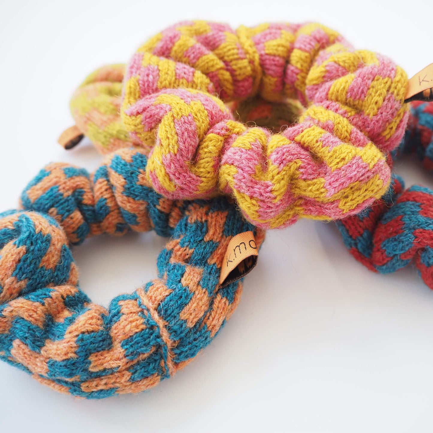 A pile of colourful knitted scrunchies with a thick zig-zag pattern, by K.Moods. They are red, yellow, blue, lime, pink, and orange.