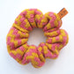 A knitted scrunchie with a thick zig-zag pattern by K.Moods. It is pink and yellow, and is on a white background.