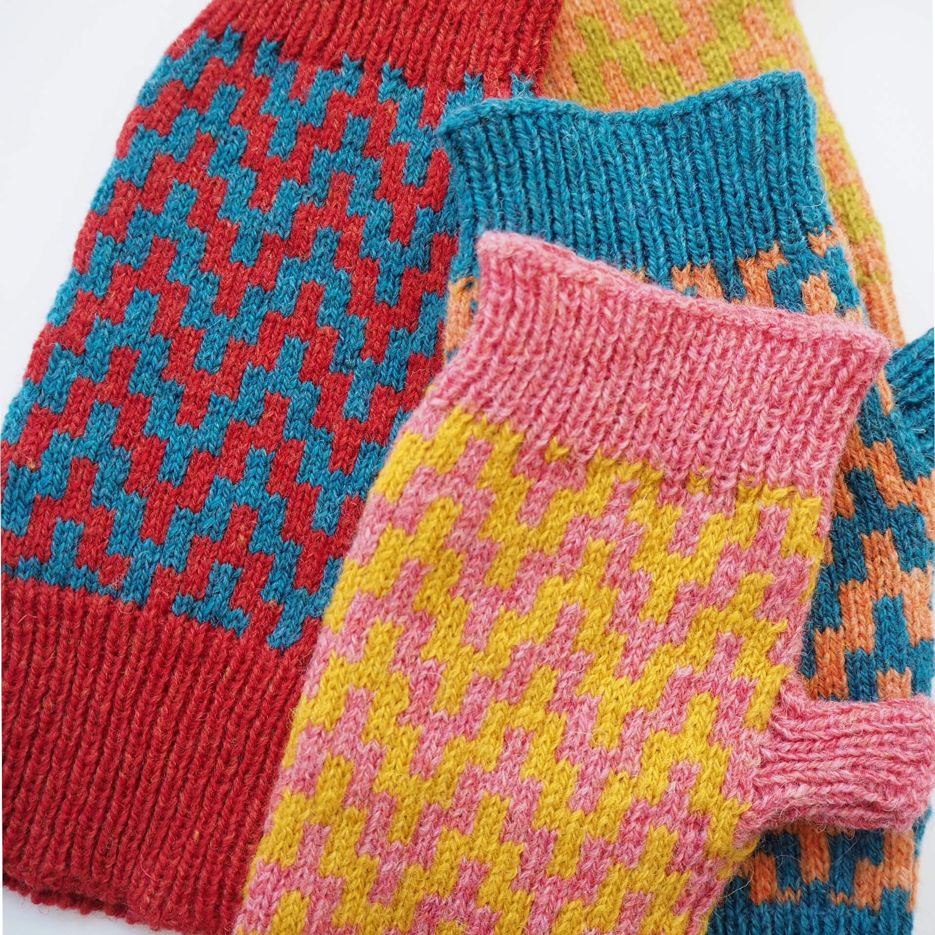 A pile of fingerless gloves by K.Moods. They have a thick zig-zag pattern, and are red, blue, yellow, pink, orange, and lime green.