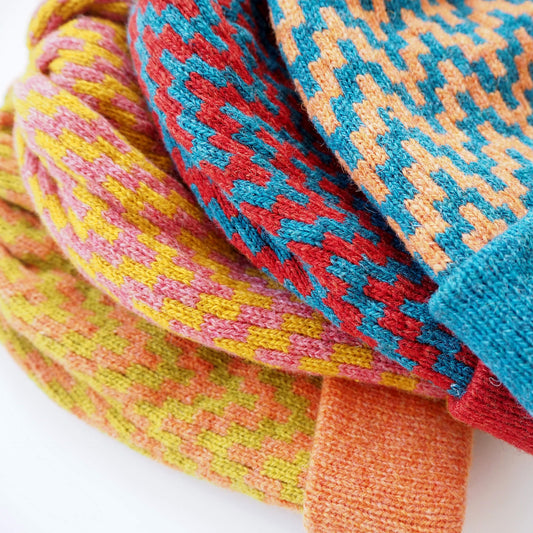 A pile of four colourful beanies on top of each other. The beanies have a zig-zag pattern, and fold up brims. They are orange, red, lime green, blue, yellow, and pink.