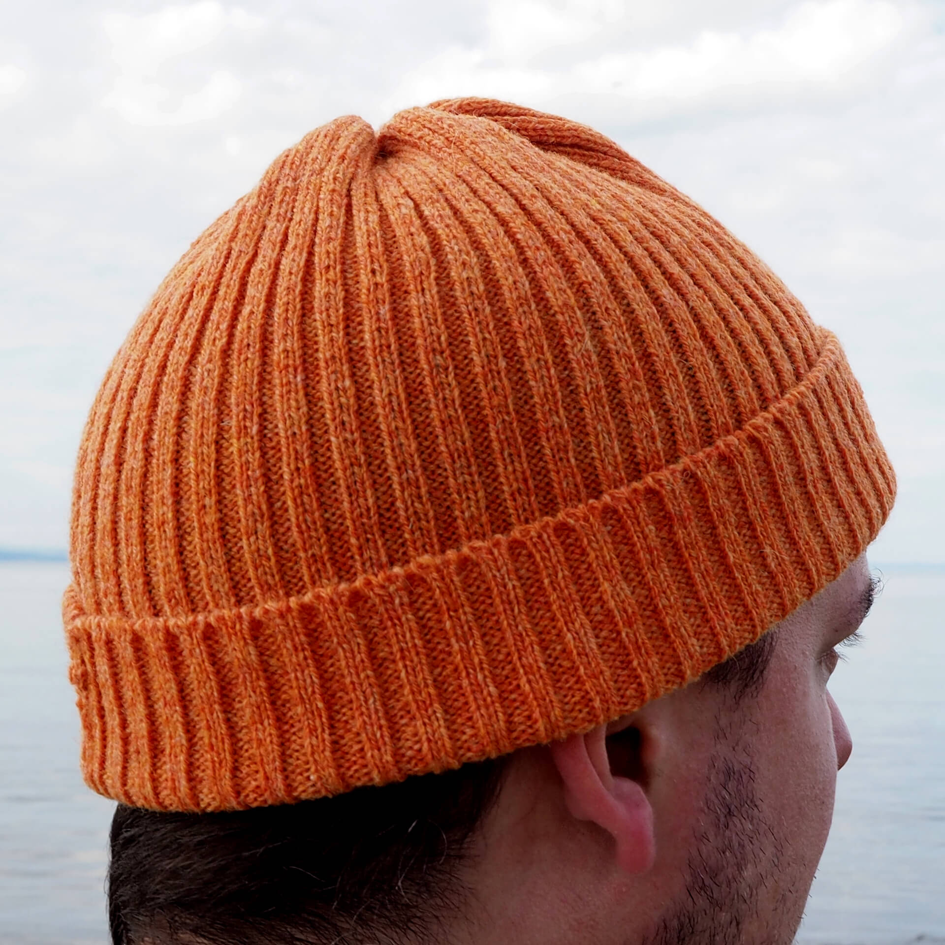 A man is wearing an orange ribbed beanie by K.Moods. He is looking away from the camera.