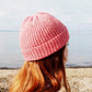 A woman wearing a pink ribbed beanie with a folded brim looks into the distance. 
