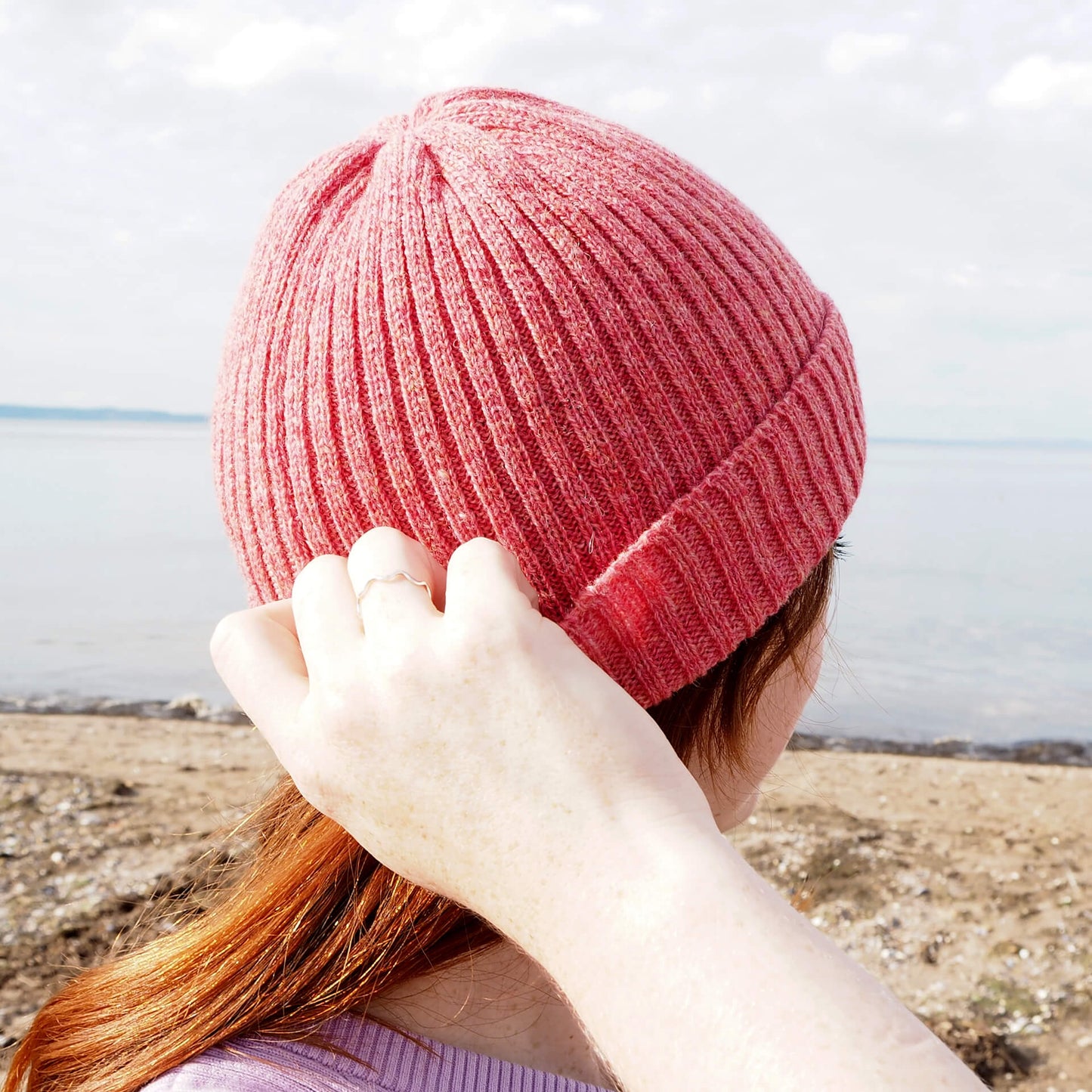 A woman adjusts a pink ribbed beanie as she looks into the distance away from the camera.