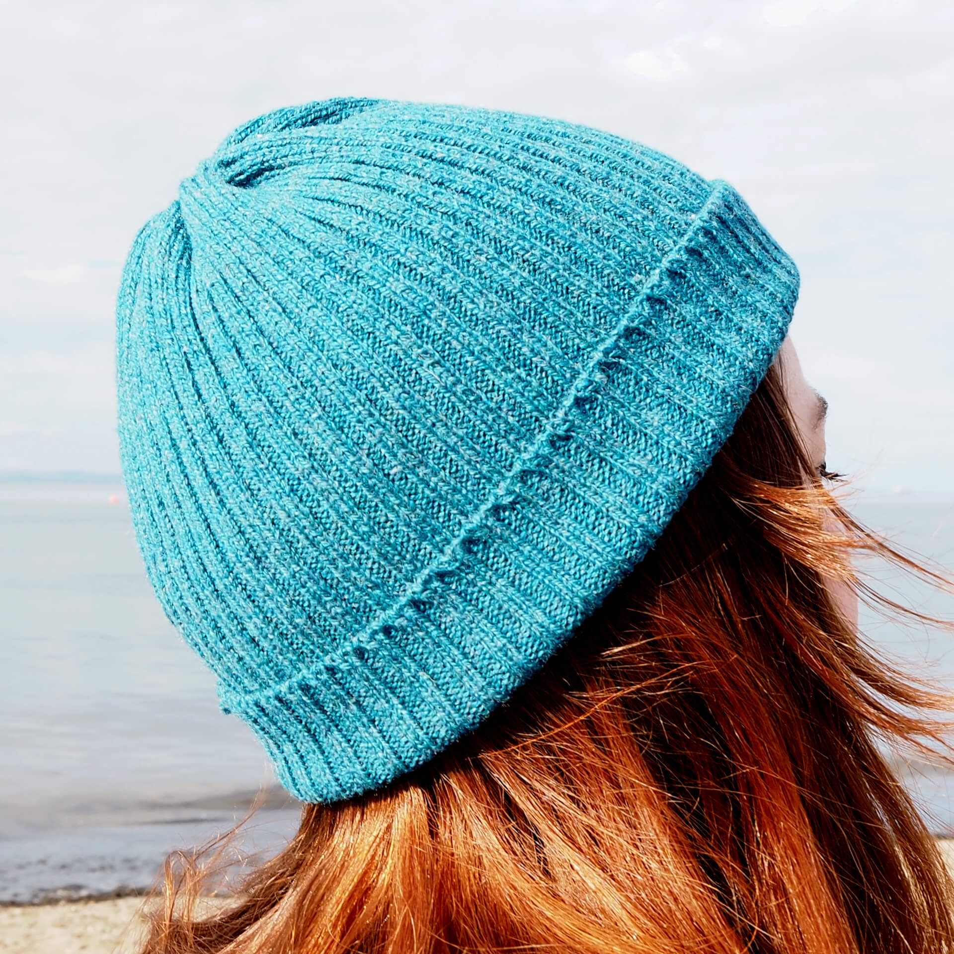 A view from the side of a woman with red hair wears a blue ribbed beanie by K.Moods.