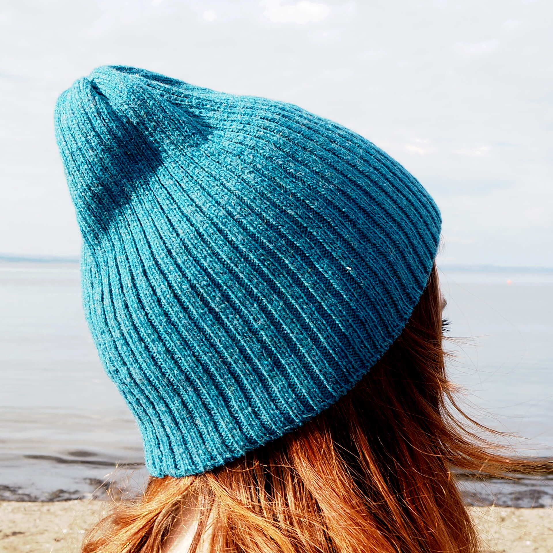 A woman with red hair wears a blue ribbed beanie by K.Moods. The beanie is slouchy at the top.