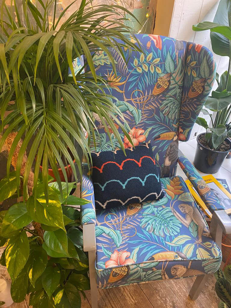 A colourful wingback chair sits surrounded by leafy green houseplants. A knitted navy cushion with a rainbow scallop pattern adorns the chair.