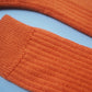 A pair of socks by K.Moods. Knitted with bright orange yarn, a thick cuff, and repetitive ribbing around the leg and on the top of the foot. This picture shows a close up of the ribbing.