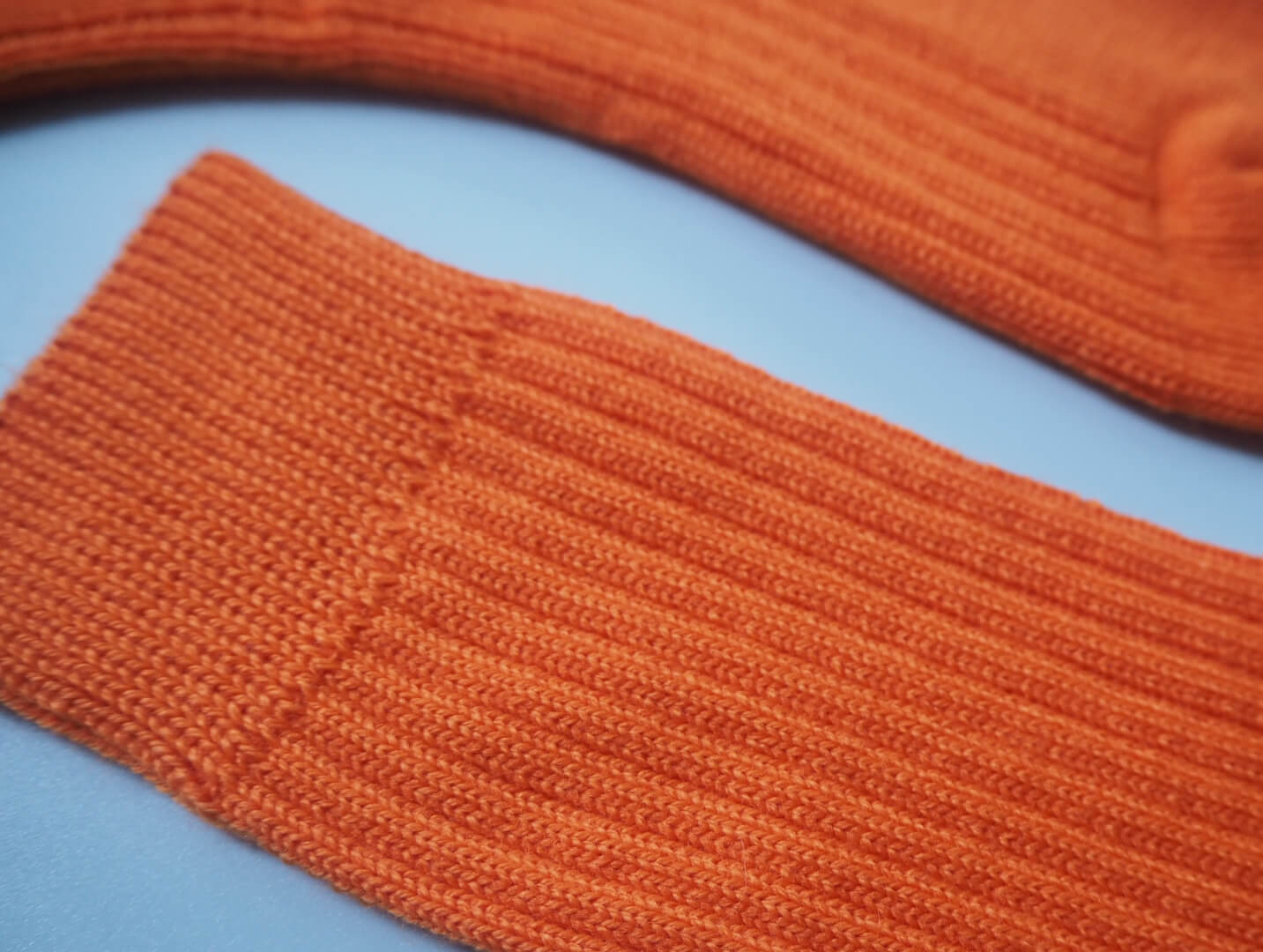 A pair of socks by K.Moods. Knitted with bright orange yarn, a thick cuff, and repetitive ribbing around the leg and on the top of the foot. This picture shows a close up of the ribbing.