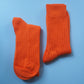 A pair of socks by K.Moods. Knitted with bright orange yarn, a thick cuff, and repetitive ribbing around the leg and on the top of the foot. This picture shows the socks folded in half, sitting side by side.