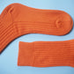 A pair of socks by K.Moods. Knitted with bright orange yarn, a thick cuff, and repetitive ribbing around the leg and on the top of the foot. This picture shows the socks lying flat with a focus on the foot and the leg.