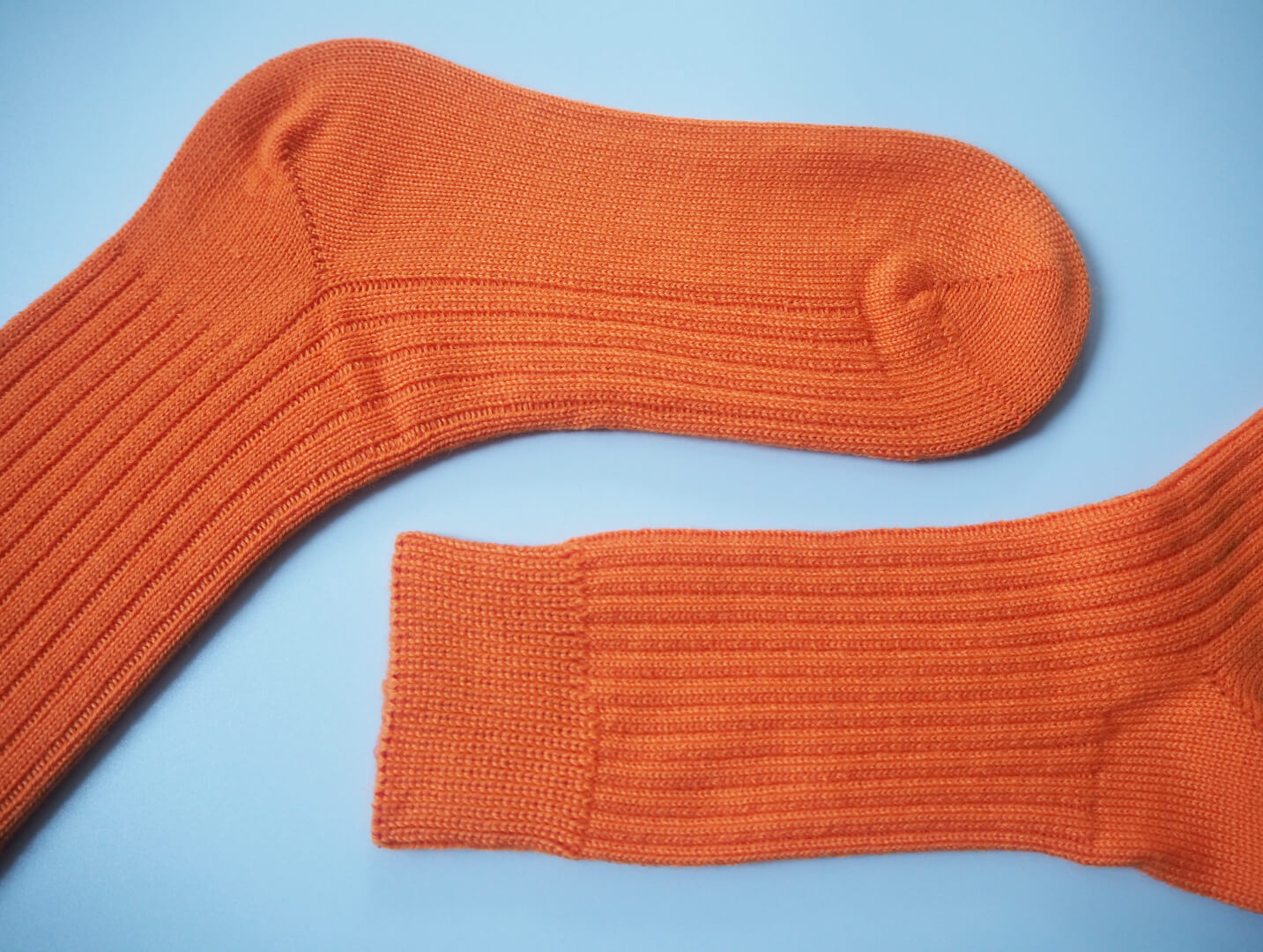 A pair of socks by K.Moods. Knitted with bright orange yarn, a thick cuff, and repetitive ribbing around the leg and on the top of the foot. This picture shows the socks lying flat with a focus on the foot and the leg.